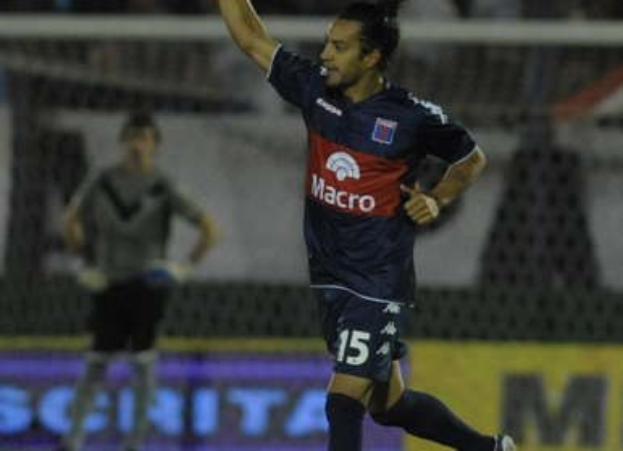 Ein Fußballspieler feiert auf dem Boden nach einem Tor, mit einer jubelnden Menge und einer Person im Hintergrund und einer Tafel mit Text dahinter, alles vor einem leicht unscharfen, energetischen Hintergrund.