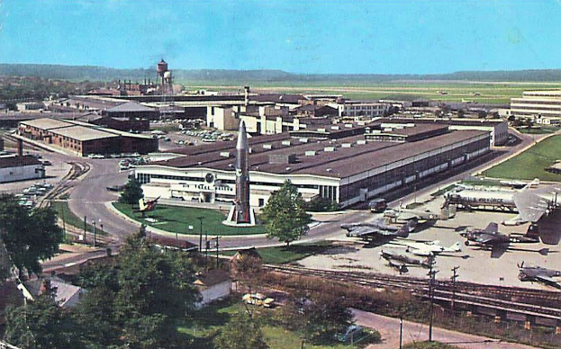 Alte Schwarz-Weiß-Airport-Szene mit geparkten Propellerflugzeugen, Terminalgebäuden, Straßen, Fahrzeugen, Versorgungsmasten, Grasflächen, Bahnschienen, fernen Hügeln und einem klaren Himmel; Bildunterschrift erscheint unten.