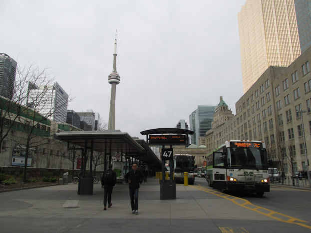 Eine belebte Stadtstraße mit einem Bus an einer Bushaltestelle, Fahrzeuge, Fußgänger, eine Hütte, Bäume, hohe Gebäude, ein Turm und der Himmel.