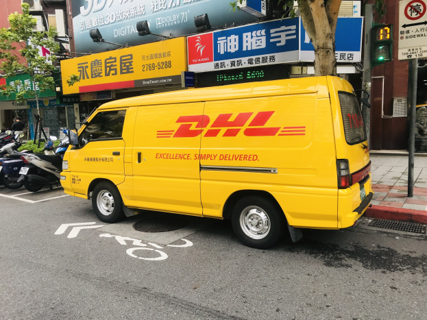Gelber DHL-Lieferwagen auf der Straßenseite geparkt, umgeben von Gebäuden, Bäumen, Schildern, Mästen, Verkehrszeichen und einer Person auf dem Gehweg.