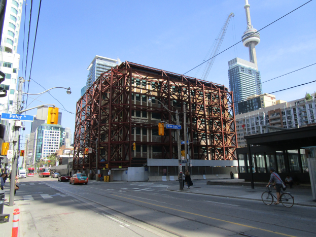 Eine belebte Stadtstraße mit einem großen Baugerüst im Hintergrund, mit Fahrzeugen, einem Radfahrer, Fußgängern, Ampeln, Pfählen, Schildern, einem Kran und Gerüsten.
