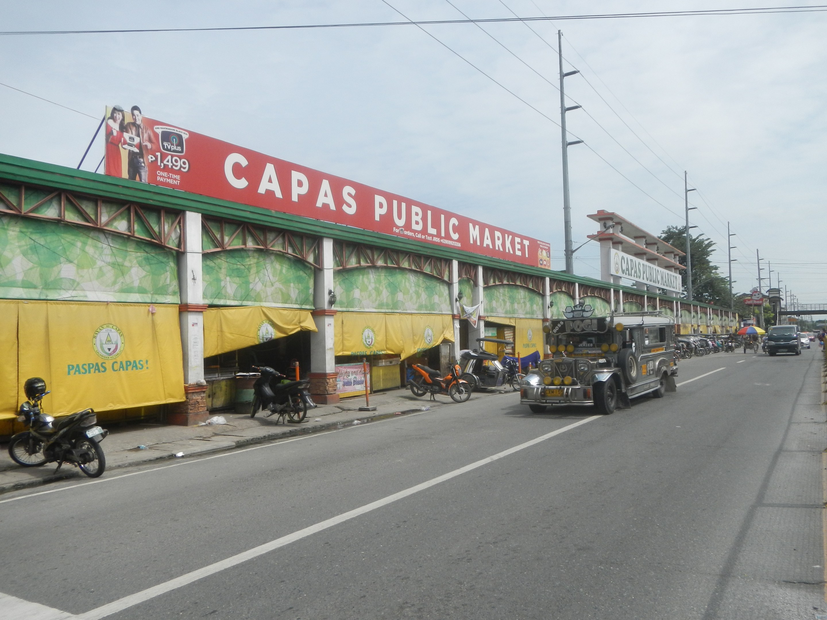 Eine belebte Straßenansicht mit Fahrzeugen, einem Gehweg, Strommasten, Gebäuden, Bäumen und einem bewölkten Himmel, mit einem Gebäude mit der Aufschrift "Capas Public Market" im Vordergrund.