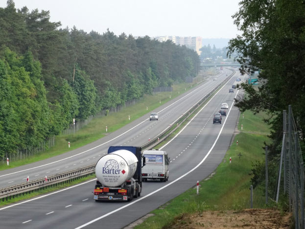 Ein großer Lkw fährt auf einer von beiden Seiten von Bäumen gesäumten Autobahn, mit einem Zaun und Gras auf der rechten Seite, Gebäuden in der Ferne und einem klaren blauen Himmel.