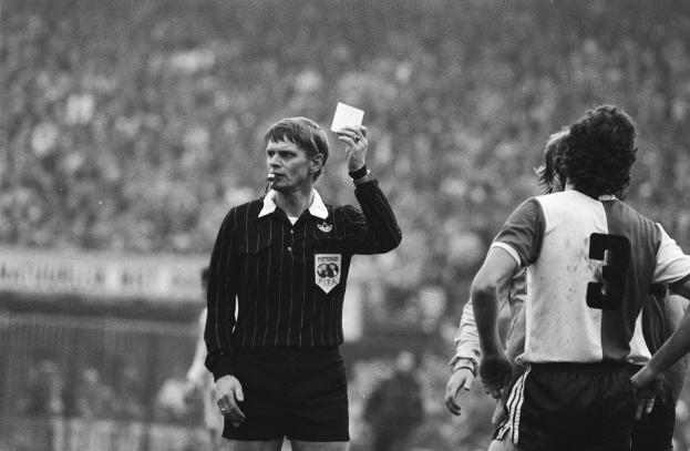 Ein Schiedsrichter in Schwarz-Weiß-Bild hält eine rote Karte vor einer Menge von Menschen in Fußballtrikots hoch, mit einem Zaun im Hintergrund.