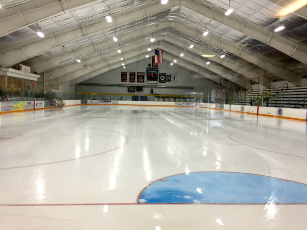 Große Indoor-Eisfläche mit Deckenbeleuchtung, Werbetafeln, einer Rückwand, Treppen mit Geländern und einigen Menschen, die auf eine Sportanlage hinweisen, wahrscheinlich eine Eishockeyhalle.