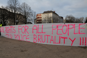 Eine Gruppe von Menschen hält ein Transparent mit der Aufschrift 'Rechte für alle Menschen Stoppt Polizeigewalt' auf dem Boden stehend mit einer Straßenlaterne, einem Schild, Bäumen, Gebäuden mit Fenstern und einem bewölkten Himmel im Hintergrund.