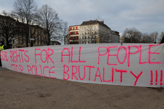 Eine Gruppe von Menschen hält ein Transparent mit der Aufschrift 'Rechte für alle Menschen Stoppt Polizeigewalt' auf dem Boden stehend mit einer Straßenlaterne, einem Schild, Bäumen, Gebäuden mit Fenstern und einem bewölkten Himmel im Hintergrund.