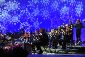 Eine Gruppe von Musikern, die auf der Bühne mit Mikrofonen, Blumengestecken und einem Bildschirm mit Schneeflocken im Hintergrund spielen.