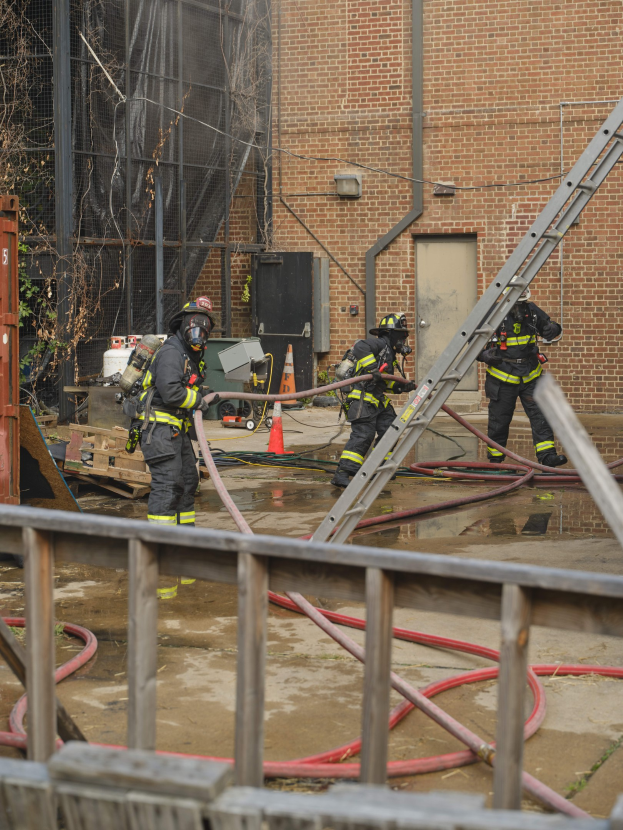 Feuerwehrleute in Helmen arbeiten daran, ein Gebäude Feuer zu löschen, während sie Schläuche halten, mit einem Metallzaun, verstreuten Rohren, einem Container, einer Verkehrskegel und Hintergrundelementen wie einem Baum und Himmel.