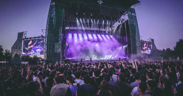 Eine große Menschenmenge auf einem Musikfestival schaut auf der hell erleuchteten Bühne mit Instrumenten und Bildschirmen, die vor einem Gebäude, Bäumen und einem klaren blauen Himmel liegen.