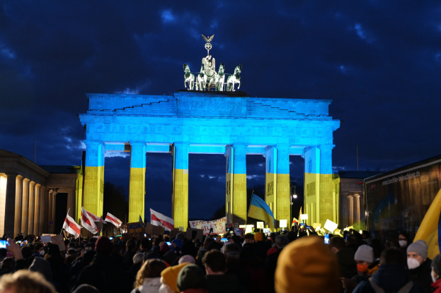 Menschenmenge mit Fahnen und Schildern vor dem Brandenburger Tor in Berlin, mit einer Fahne auf der rechten Seite.