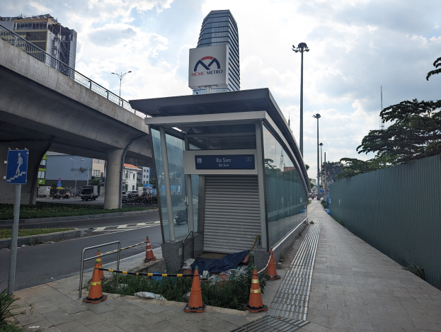 Busbahnhof mit Verkehrskegeln, Pflanzen und einem Schild neben einem hohen Gebäude, umgeben von Fahrzeugen, einer Brücke und einem bewölkten Himmel.