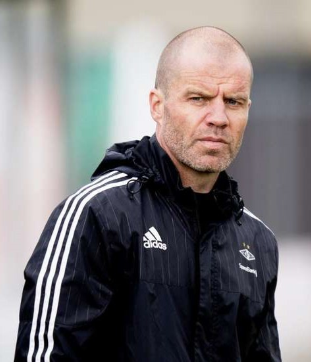 Baldiger männlicher Fussballtrainer in marineblauer Jacke mit weißen Streifen und weißem Kragen, der auf einem Fussballfeld steht und nachdenklich schaut.