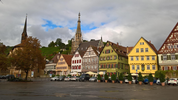 Ein idyllischer Marktplatz in Rothenburg ob der Tauber, Deutschland, mit Gebäuden mit Fenstern, Topfpflanzen, Bäumen, Fahrzeugen, Schildern und einer Kirche im Hintergrund bei bewölktem Himmel.