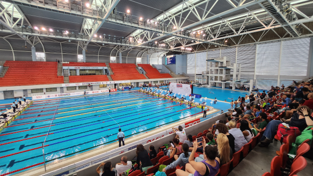 Großer Hallenbad mit vielen Menschen auf Stühlen drumherum und einigen im Wasser Stehenden, mit hoher Decke mit Lampen und Eisenstangen und Hintergrundtafeln mit Text, die das Olympische Schwimmzentrum in London anzeigen.