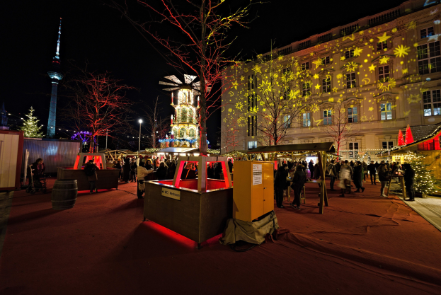Ein belebter Weihnachtsmarkt in Berlin, Deutschland, mit Menschen um geschmückte Stände, festliche Lichter, Bäume, Gebäude, Laternenmasten und einen Turm unter einem dunklen Himmel.