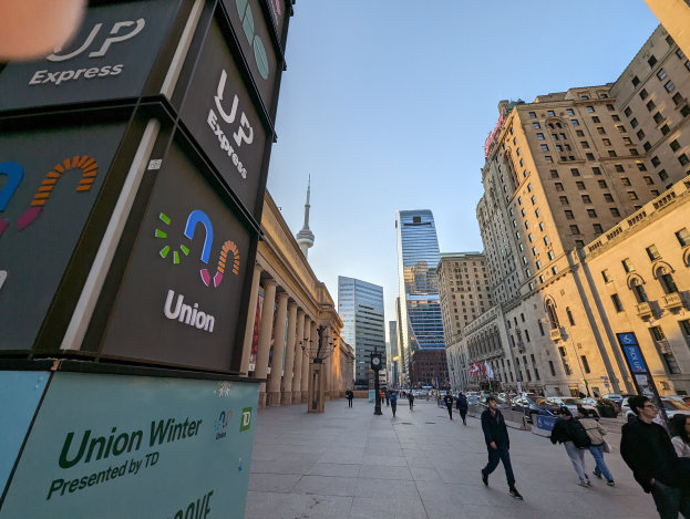 Eine belebte Stadtstraße mit Fußgängern, hohen Gebäuden, Fahrzeugen und Texttafeln auf der linken Seite, unter einem sichtbaren Himmel.