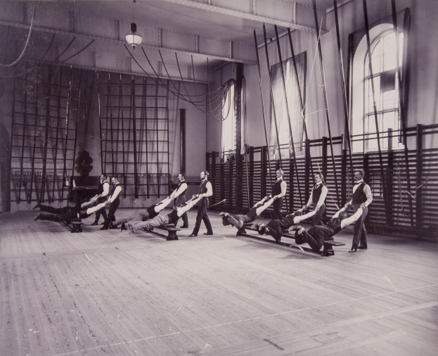 Schwarzes und weißes Bild einer Gruppe von Menschen, die Pilates-Übungen in einem Fitnessstudio mit einer Wand im Hintergrund, Fenstern auf der rechten Seite und Deckenbeleuchtung durchführen.