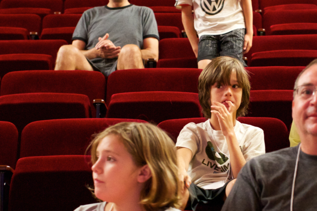 Eine Gruppe von Menschen in einem mit roten Sesseln gefüllten Kino, mit einem Jungen in einem weißen T-Shirt und schwarzen Hosen in der Mitte.