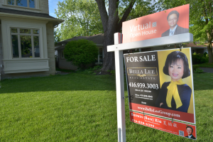 Ein Immobilien-Verkaufsschild steht vor einem Haus mit sichtbaren Fenstern, umgeben von saftigem Gras, Bäumen und Pflanzen unter einem klaren Himmel.