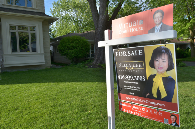 Ein Immobilien-Verkaufsschild steht vor einem Haus mit sichtbaren Fenstern, umgeben von saftigem Gras, Bäumen und Pflanzen unter einem klaren Himmel.