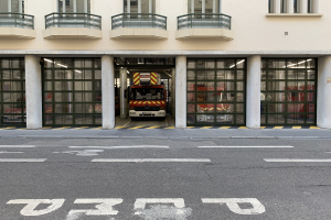 Eine Feuerwache mit einem Feuerwehrauto davor, das Fenster, Geländer, Säulen und Glastüren zeigt, mit einer Straße unten.