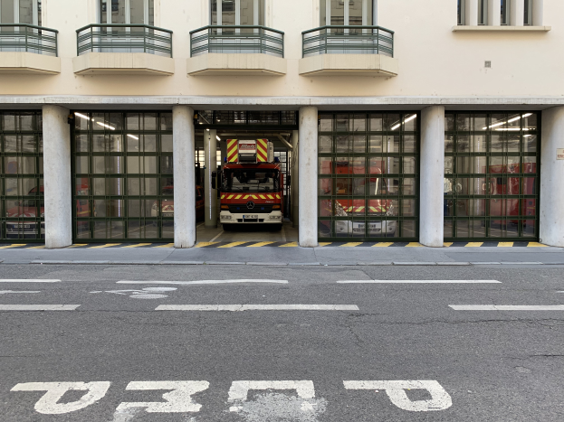 Eine Feuerwache mit einem Feuerwehrauto davor, das Fenster, Geländer, Säulen und Glastüren zeigt, mit einer Straße unten.