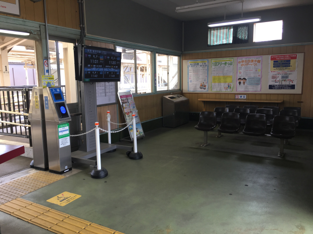 Ein belebter Bahnhofseingang mit Sitzgelegenheiten, einem Servicecounter, Ticketautomaten, Deckenleuchten, Geländern, Informationsanzeigen und Wandplakaten.