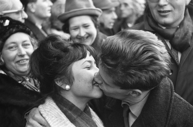 Schwarzes und weißes Foto eines Mannes und einer Frau, die leidenschaftlich vor einer lachenden Menge knutschen.