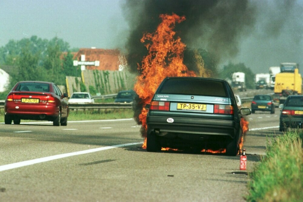 Ein Auto, das in Flammen steht, am Straßenrand mit anderen Fahrzeugen in der Nähe, Bäumen und Gebäuden im Hintergrund, einem klaren blauen Himmel, Gras auf der rechten Seite und einem Feuerlöscher.