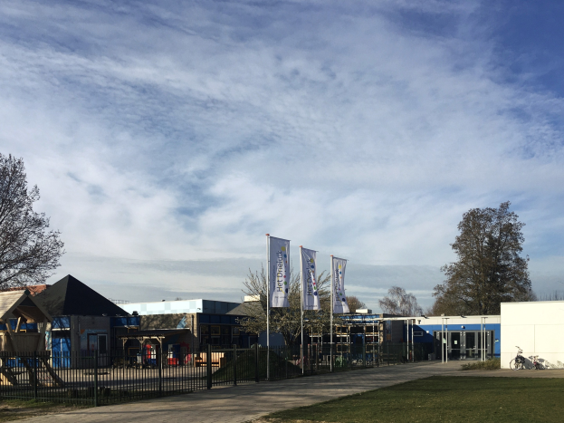 Großes Gebäude mit einem Metallzaun, Rasenfläche, Weg, Bäumen, Bannern, einem geparkten Fahrrad, nahen Häusern und einem bewölkten Himmel, identifiziert als der neue Spielplatz an der Schule.