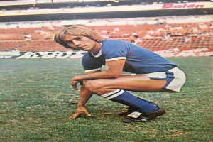 Ein Mann kniet auf einem Fußballfeld, umgeben von sitzenden Menschen mit Stühlen und Tafeln mit Text.