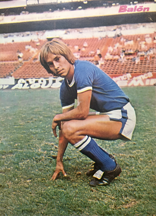 Ein Mann kniet auf einem Fußballfeld, umgeben von sitzenden Menschen mit Stühlen und Tafeln mit Text.