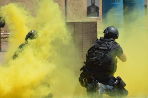 Zwei Polizisten in Schutzausrüstung mit gelbem Rauch aus dem Mund, mit Helmen und Schutzausrüstung vor einer Wand mit einer Tafel und anderen Gegenständen, mit sichtbarem Boden.