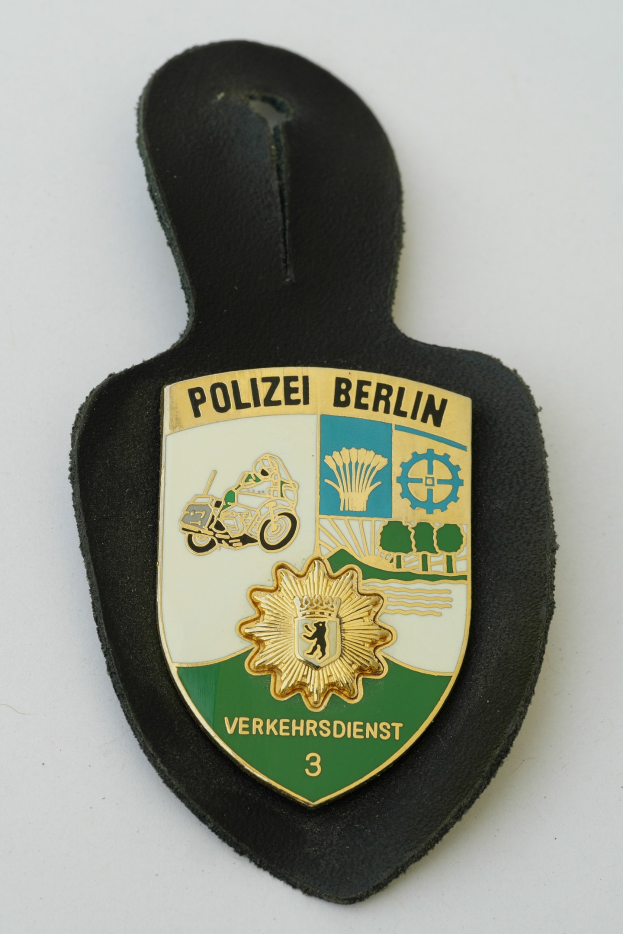 Ein schildförmiges Polizeibadge mit 'Polizei Berlin' in schwarzer fettgedruckter Schrift auf einem blauen Hintergrund mit einem weißen Stern und Rahmen, das auf einer Oberfläche ruht.