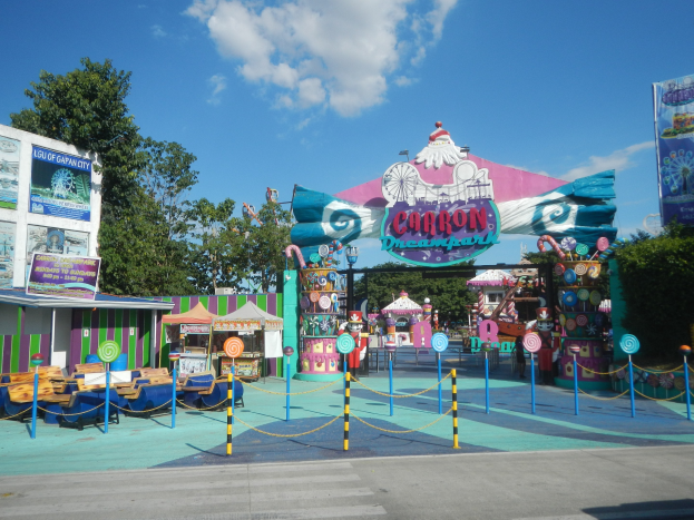 Kirmesfahrgeschäft am Parkeingang mit Straße darunter, eingezäuntes Gebiet, Buden, Banner, Bäume und bewölkter Himmel im Hintergrund.