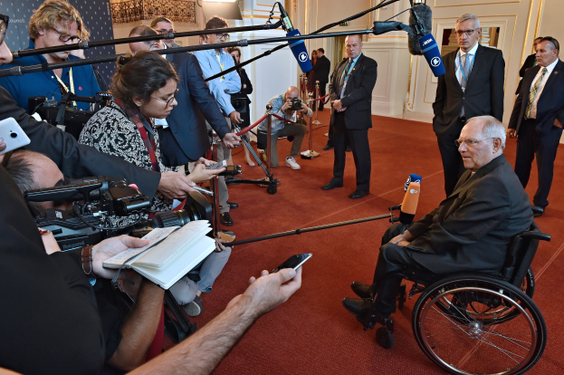 Ein Mann im Rollstuhl spricht mit Reportern, die Kameras, Mikrofone und Handys halten, vor einem Banner, Scheinwerfern und einer Wand.