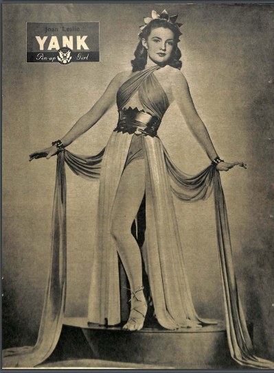 Ein Schwarz-Weiß-Foto von Schauspielerin Joan Leslie in einem langen, hochgeschlossenen Kleid mit langen Ärmeln, die selbstbewusst auf einer Bühne steht.