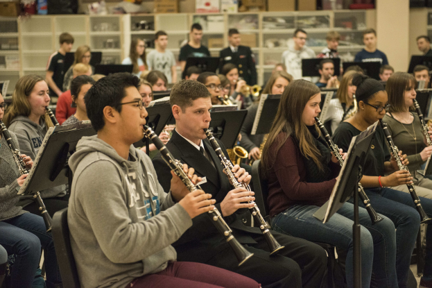 Eine Gruppe von Menschen, die Klarinetten in einem Klassenzimmer spielen, einige sitzen mit Notenpulten und andere stehen, mit Regalen voller Kisten im Hintergrund.