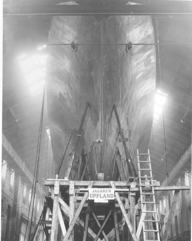 Schwarzes und weißes Foto eines großen Boots im Bau in einer Werft, das eine Leiter, Holzpfosten, ein Schild, Metallrahmen, Deckenlampen und Fenster zeigt.