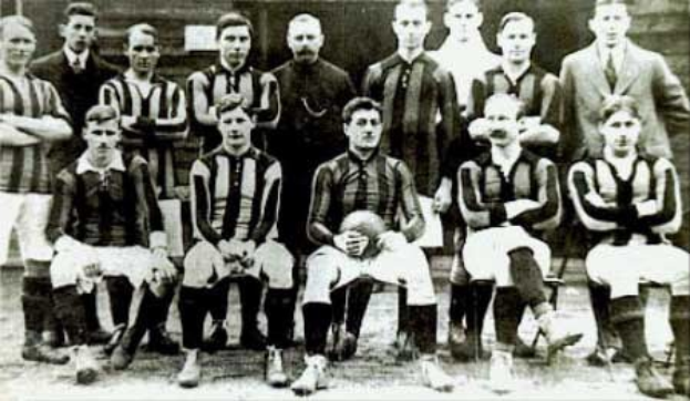 Schwarzes und weißes Foto einer Fußballmannschaft, einige stehen und einige sitzen, vor einer Wand mit dem Text «Ballymena Football Club» unten.