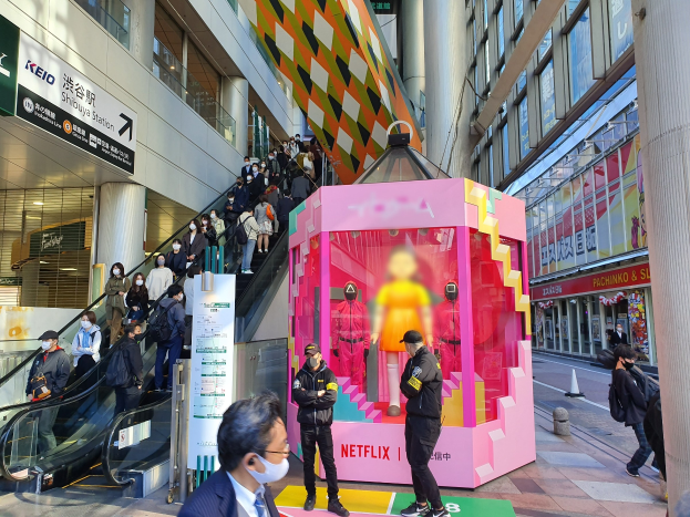 Eine Gruppe von Menschen steht vor einem Gebäude mit Glaswänden und Texttafeln, einige tragen Masken, in der Nähe einer Rolltreppe; ein pinker Kasten in der Mitte zeigt Schaufensterpuppen und trägt den Text "Netflix's new Netflix booth in Tokyo."