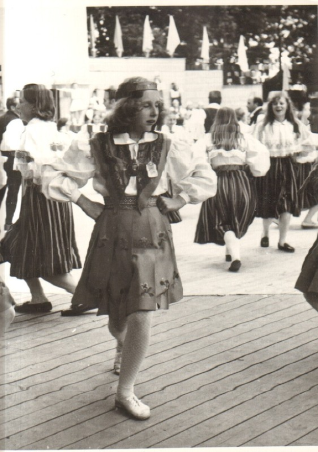 Eine Gruppe von Frauen in traditioneller bayrischer Tracht tanzt auf einem Holzboden, mit einer Wand, Bäumen und anderen Gegenständen im Hintergrund und Text am unteren Bildrand.