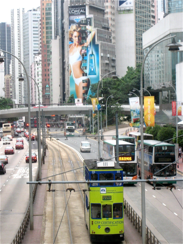 Eine belebte Stadtstraße mit Doppeldeckerbussen, Autos und anderen Fahrzeugen unter einem klaren blauen Himmel, mit hohen Gebäuden, Bäumen, Straßenlaternen, Schildern und einer Brücke im Hintergrund, mit Text unten.