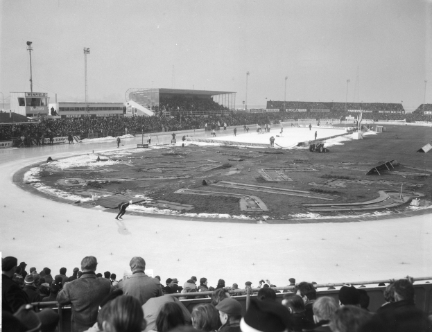 Schwarzes und weißes Foto eines Eishockeyspiels in einer Arena mit Zuschauern, Pfosten, Gebäuden und einem klaren Himmel im Hintergrund.
