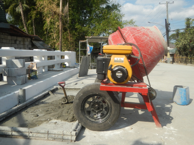 Betonmischer auf einem Zementlastwagen umgeben von Baumaterialien, einem Haus, Bäumen, Strommasten und einem bewölkten Himmel.