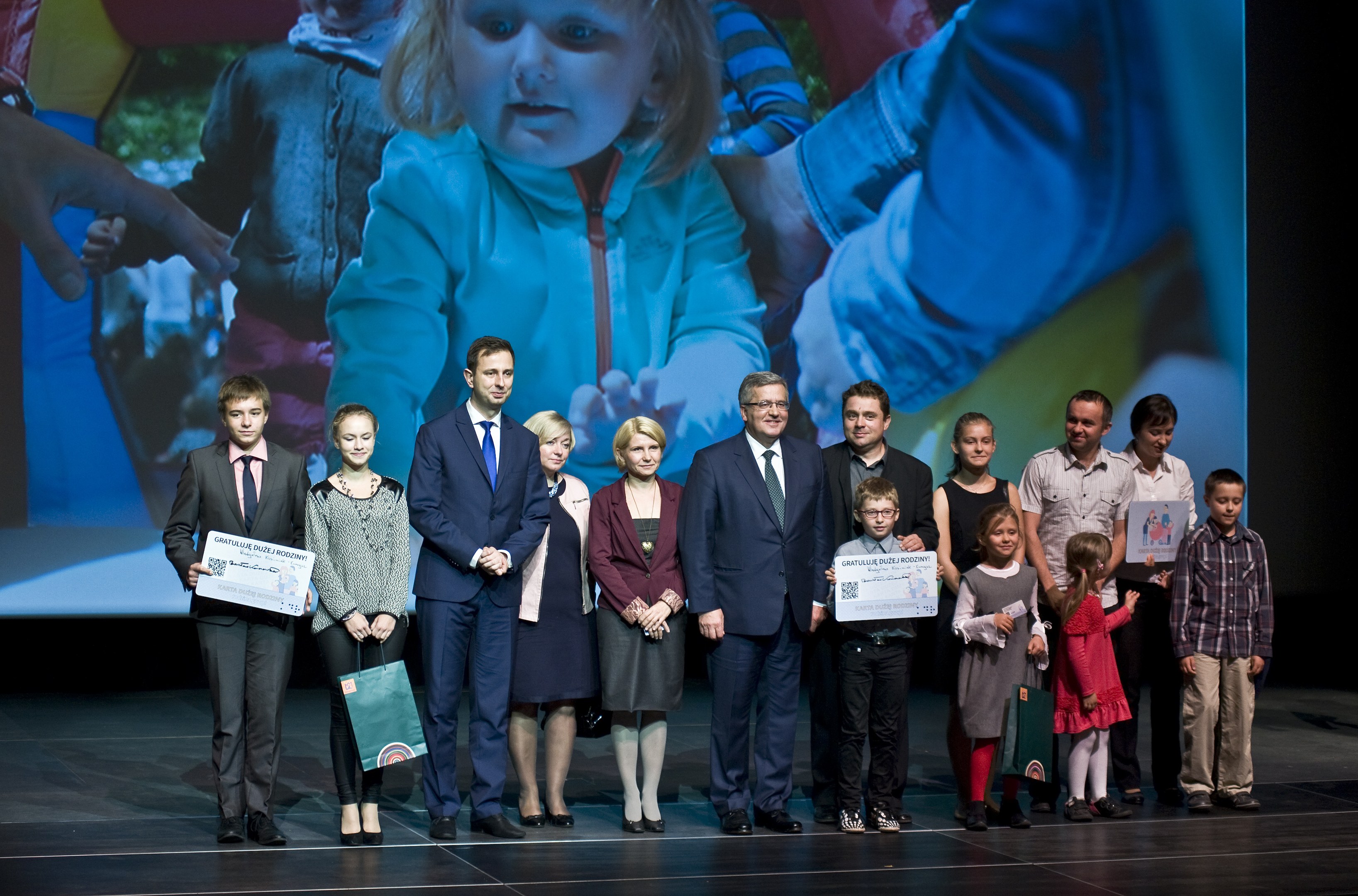 Gruppe von Menschen auf einer Bühne mit Taschen und Urkunden, im Hintergrund ein Bildschirm mit einem Kind und anderen