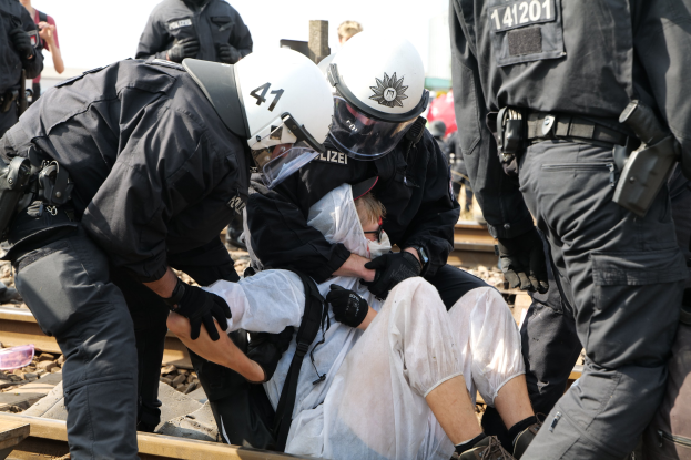 Eine Gruppe von Polizisten in Einsatzkleidung nimmt einen Mann in einem weißen Kleid auf einem Gleis fest, während andere im Hintergrund stehen.