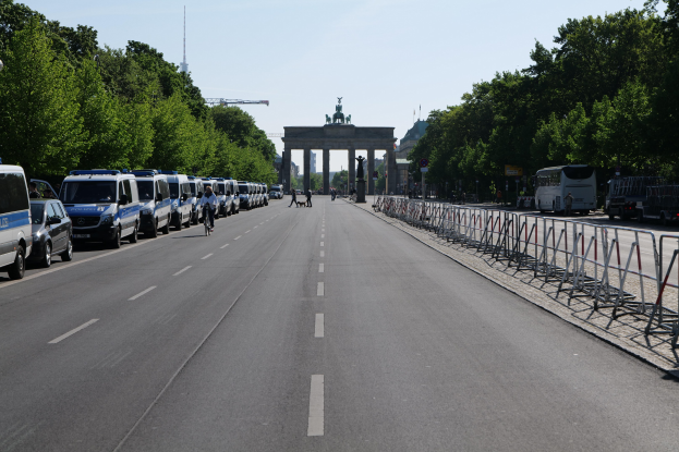 Lange Reihe von Polizeiwagen am Straßenrand vor dem Brandenburger Tor in Berlin, Deutschland, mit Menschen auf Fahrrädern und auf der Straße stehend, Barrieren, Bäume, ein Bogen mit Statuen im Hintergrund und sichtbarem Himmel.