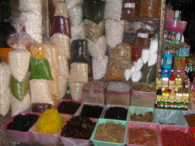 Marktstand mit einer Auswahl an Lebensmitteln in Flaschen, Schachteln und anderen Behältern, mit ein paar Menschen im Hintergrund.